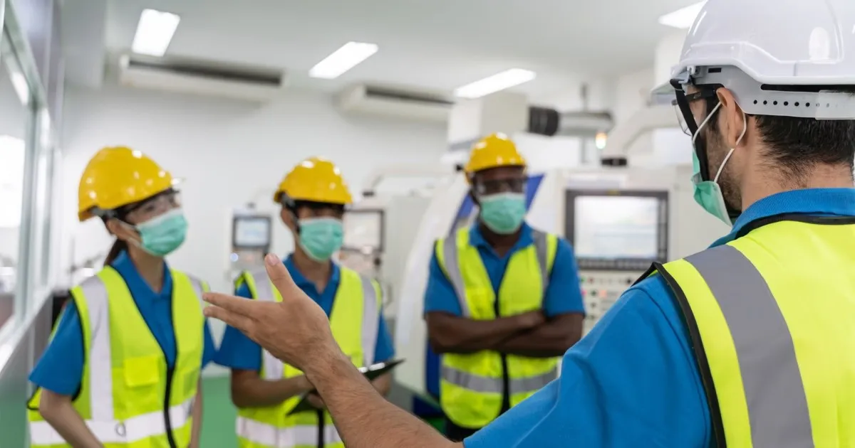 Supervisor orienta a un equipo con casco y chaleco amarillo en una línea de producción.