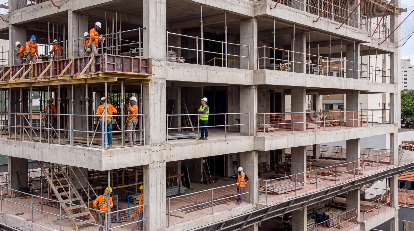 Trabalhadores da construção civil com capacete, colete e cinto de segurança em um edifício em obras, com guarda-corpos instalados nas lajes.