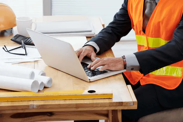 Professional in an orange high-visibility vest types on a laptop at a desk with rolled-up blueprints.