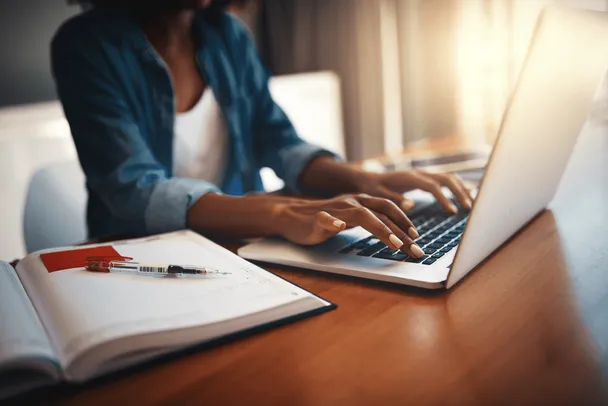 Mujer escribe en una computadora portátil junto a un cuaderno abierto con un bolígrafo.